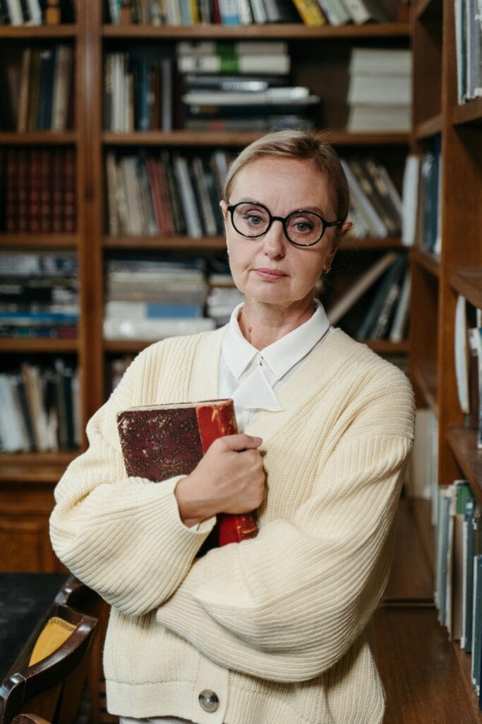 Confident woman in eyeglasses holding a book in a library setting, showcasing style and knowledge.